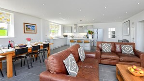 A modern kitchen with a dining area, a brown leather sofa, and a wooden table with fruit. - 5 Bed in Pwllheli (oc-dolwen) (Llanbedrog)