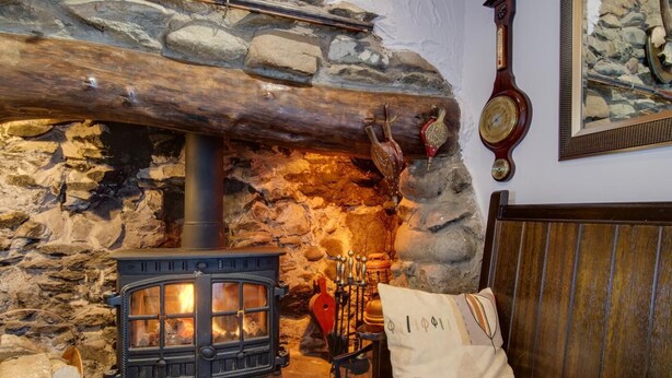 A stone fireplace with a wood-burning stove, a clock, and a bed with a red headboard. - 2 Bed in Pwllheli (oc-wag303) (Y Ffôr)