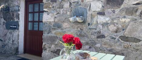 A stone building with a wooden door, a sign reading "YSGUBOR", and a table set with a vase of red flowers, two red mugs, a plate, and a loaf of bread.