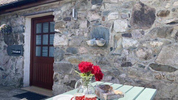 A stone building with a wooden door, a sign reading "YSGUBOR", and a table set with a vase of red flowers, two red mugs, a plate, and a loaf of bread. - 2 Bed in Y Felinheli (oc-wag431) (Y Felinheli)