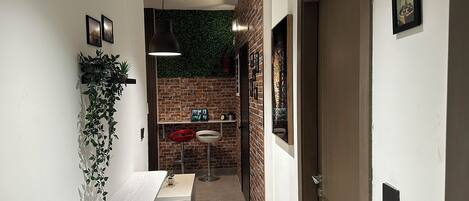 A narrow hallway with a white bench, a hanging plant, and a brick wall with a green wall hanging.