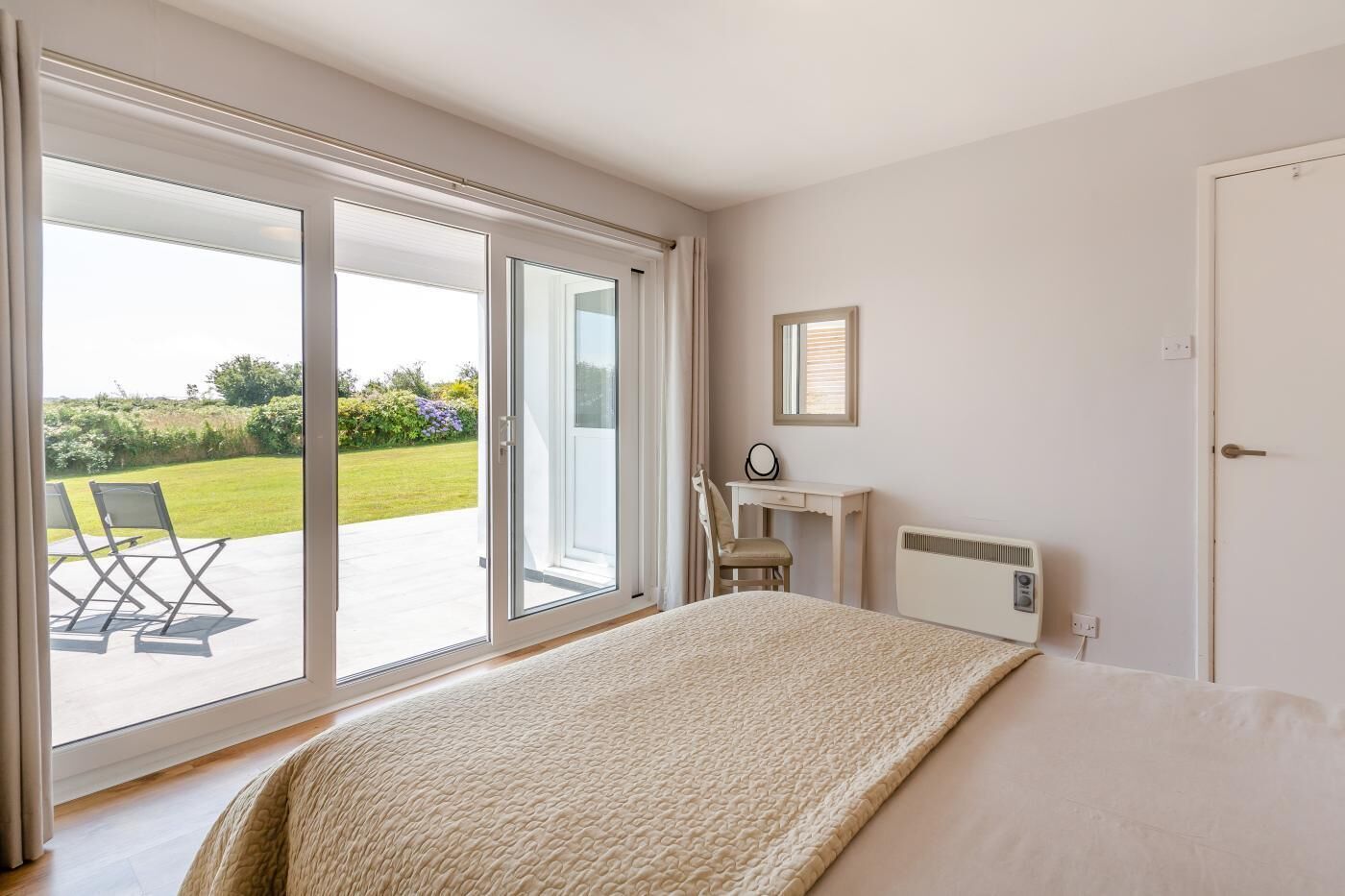 A bedroom with a large sliding glass door leading to a balcony, a bed with a textured blanket, a desk with a chair, and a radiator.