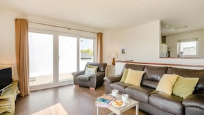A living room with a sofa, armchair, and coffee table. There is a television on a stand and a window with curtains. - 3 Bed in Abersoch (oc-4ynysf) (Abersoch)