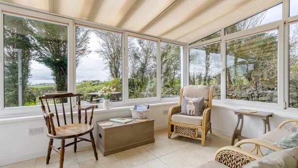 A sunlit room with large windows, a wooden chair, a wicker chair, a sofa, and a small table. - 4 Bed in Aberdaron (oc-fl055) (Aberdaron)