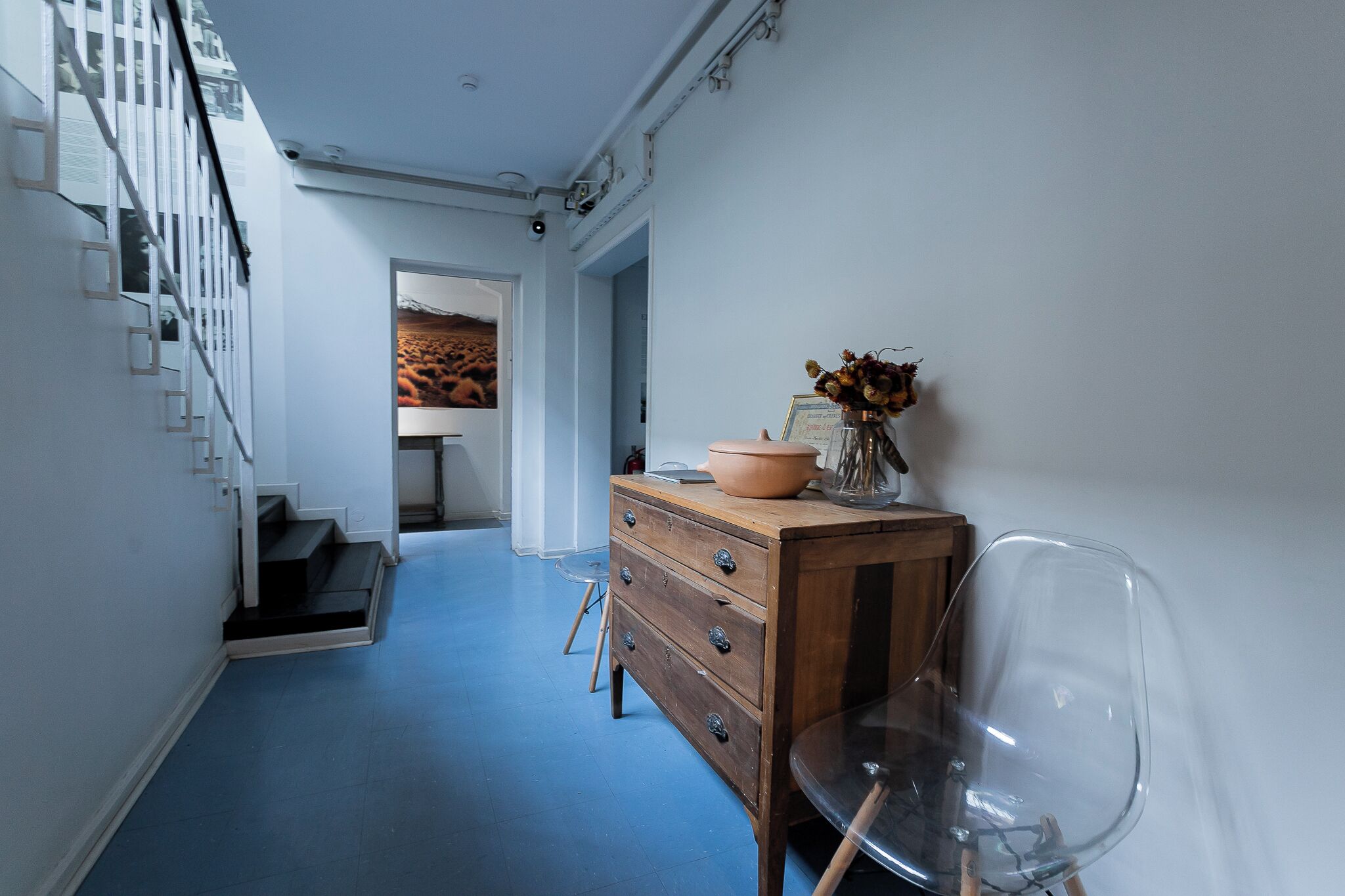 A hallway with a blue floor, a wooden dresser with drawers, a clear chair, and a staircase with white railings.