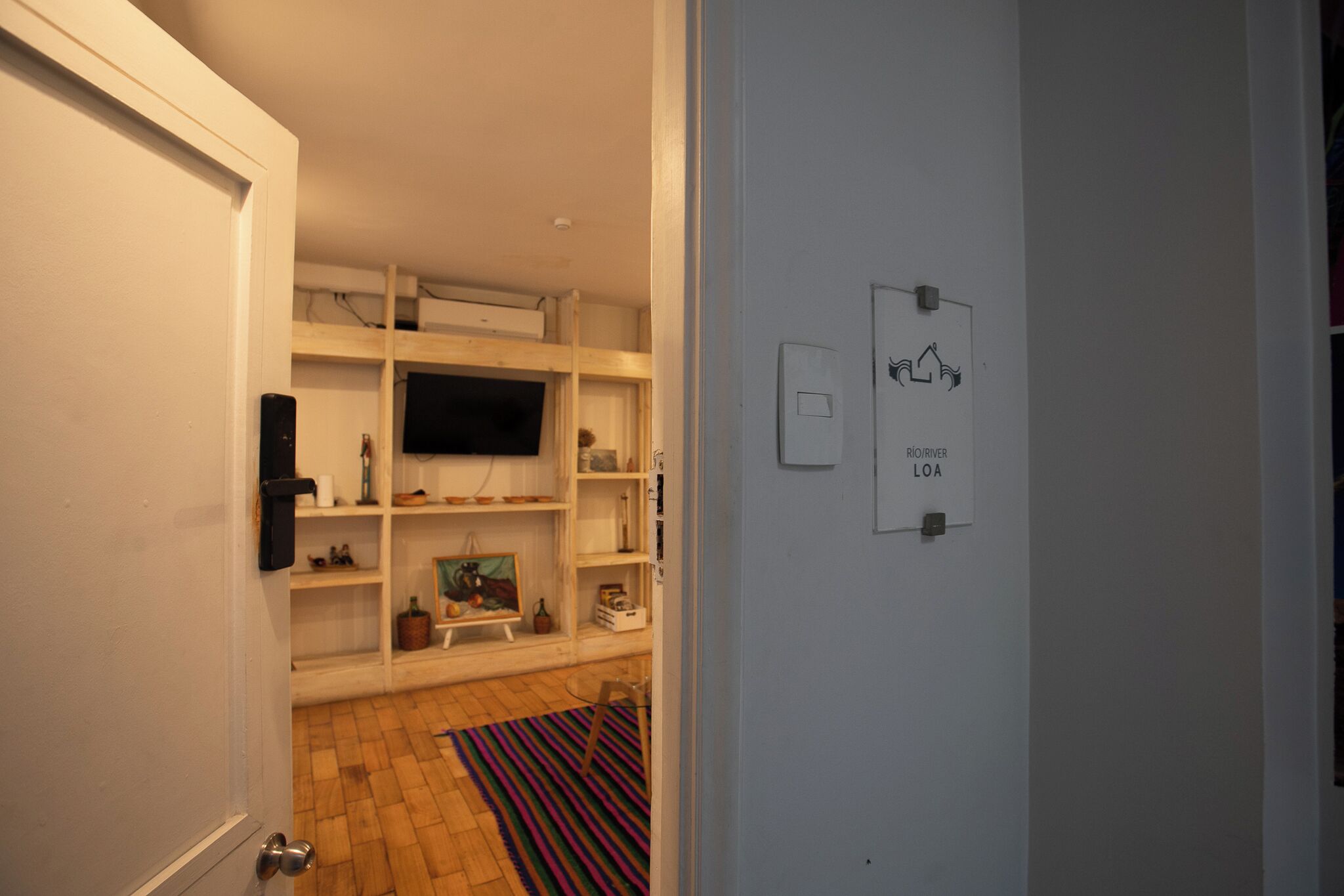 Una habitación con piso de madera, paredes blancas y un televisor montado. Hay repisas con varios objetos y un cuadro en la pared.
