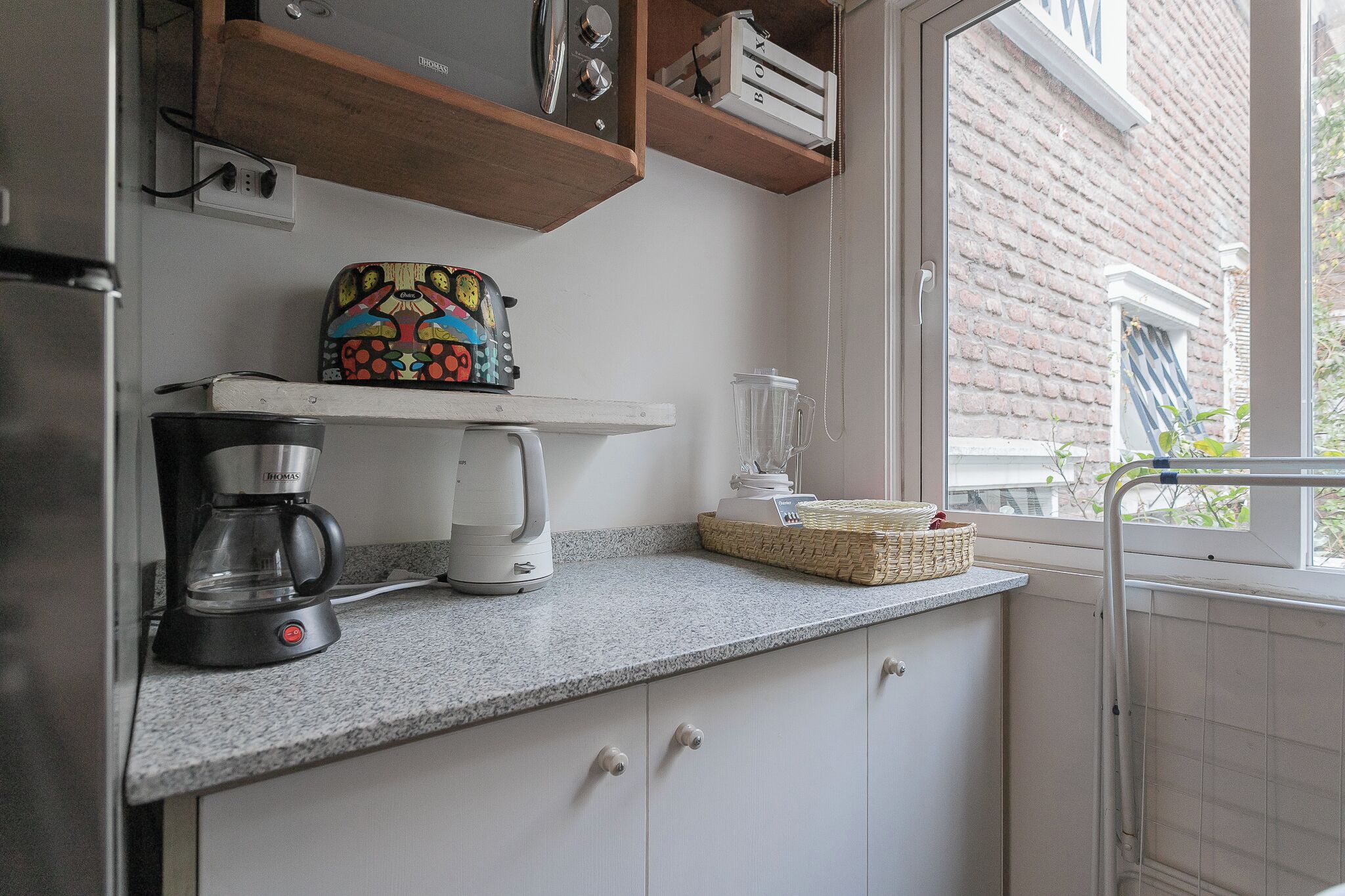 Une cuisine équipée d’une machine à café, d’un grille-pain et d’un mixeur, le tout posé sur le plan de travail. Une fenêtre offre une vue sur un bâtiment en briques situé à l’extérieur.