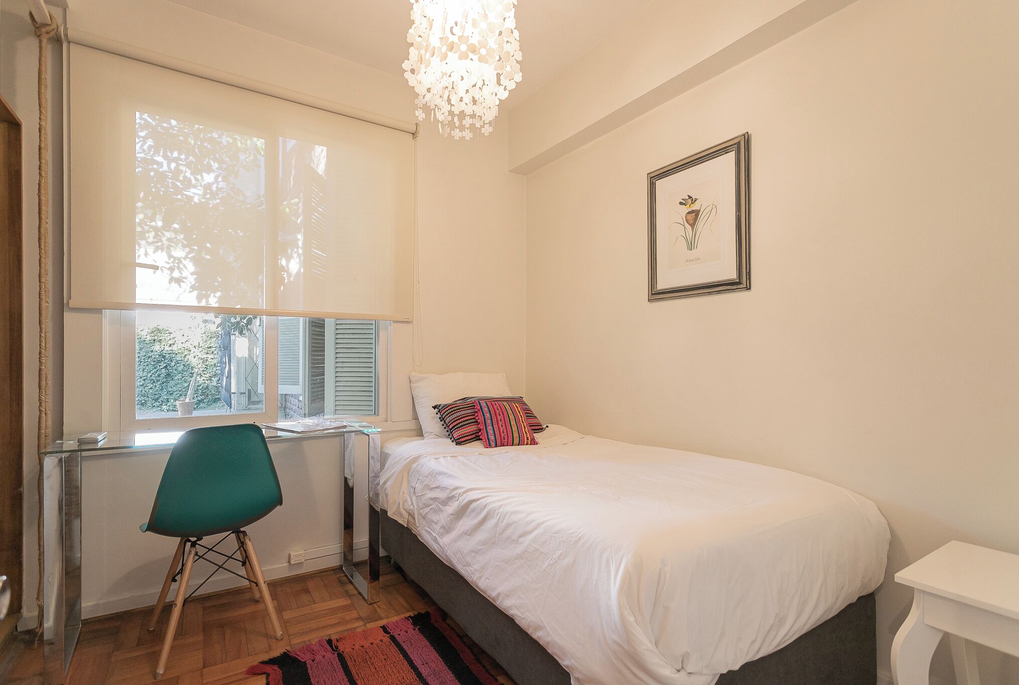 A bedroom with a bed, a desk, a chair, a window, and a chandelier.