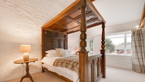 A four-poster bed with a wooden headboard and footboard, a bedside table with a lamp, and a view of the outside through a window. - 4 Bed in Pwllheli (oc-tshipp) (Abersoch)