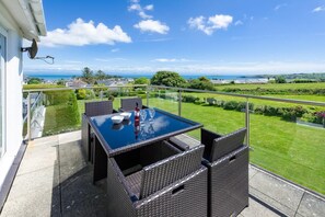 A balcony with a glass table and wicker chairs, overlooking a well-maintained lawn and distant landscape. - 3 Bed in Pwllheli (oc-5ynysf) (Abersoch)