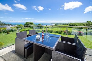 A glass-enclosed outdoor patio with wicker furniture and a table set for dining, overlooking a well-maintained garden and distant landscape. - 3 Bed in Pwllheli (oc-5ynysf) (Abersoch)