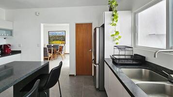 Une cuisine moderne équipée d’un réfrigérateur, d’un évier et de tabourets de bar. On aperçoit à l’arrière-plan un coin repas avec une table et des chaises.