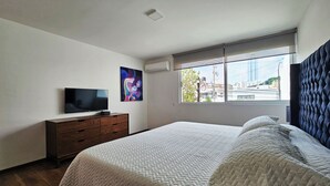 Een slaapkamer met een bed, een televisie op een dressoir, een raam met uitzicht op gebouwen en een airconditioner.