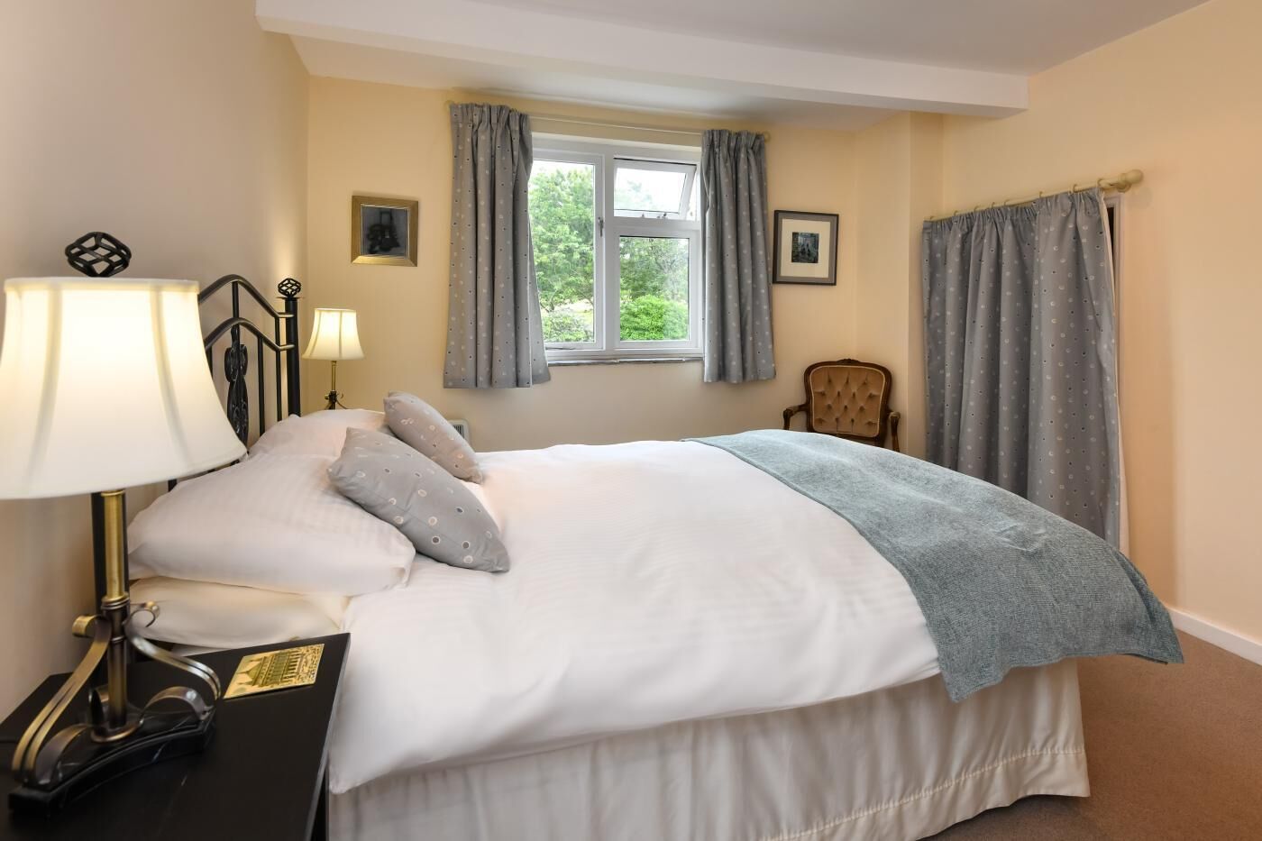 A neatly made bed with pillows and a blanket, a bedside lamp, a chair, and a window with curtains.