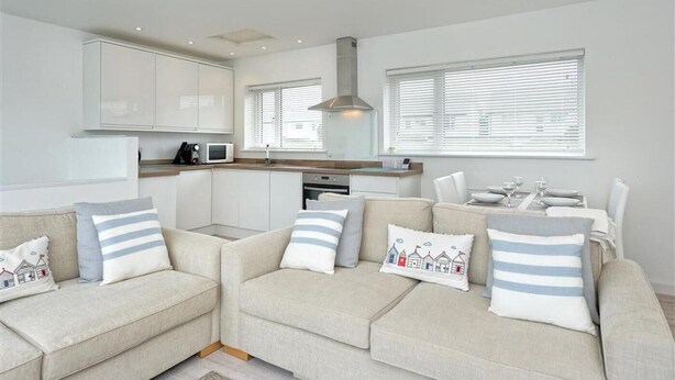 A modern living room with a beige sofa set, a kitchen area with white cabinets, and a dining table with a white tablecloth. - 2 Bed in Pwllheli (oc-45caed) (Abersoch)