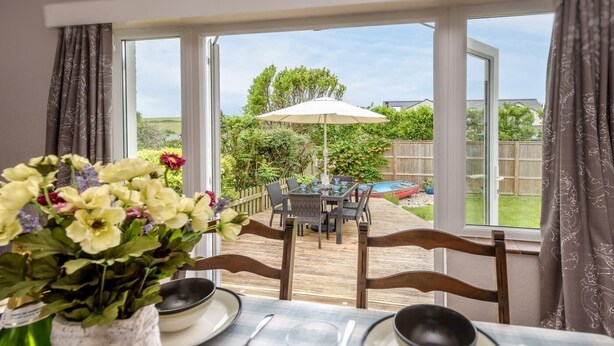 Un comedor con una mesa dispuesta para dos personas, una ventana con cortinas, y un patio exterior con mesa y sillas.
