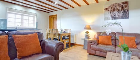 A living room with leather sofas, wooden beams on the ceiling, a dining table with chairs, and a window with blinds.