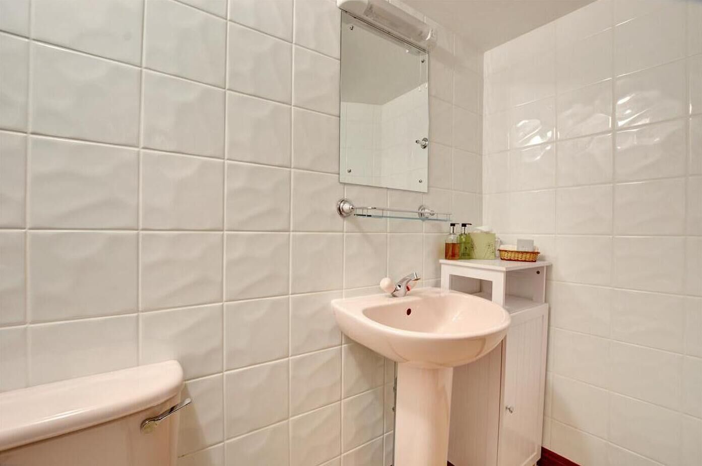 A bathroom with white tiles, a white sink, a mirror, and toiletries on a shelf.