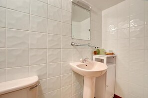 Une salle de bain avec des carreaux blancs, un lavabo blanc, un miroir et des articles de toilette rangés sur une étagère.