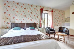 Une chambre d’hôtel avec un grand lit, deux tables de chevet, une chaise, une petite table ronde et un papier peint à motifs floraux.
