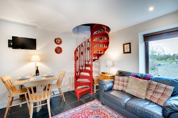 A modern living room with a spiral staircase, a dining area with wooden chairs, a sofa with plaid cushions, and a TV mounted on the wall.