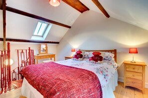 A bedroom with a sloped ceiling, a skylight, a large bed with floral bedding, two bedside tables with lamps, and a wooden dresser.
