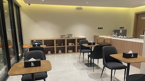 Een modern café met zwarte stoelen en houten tafels, een balie met koffiemachines en planken met kommen en kopjes.