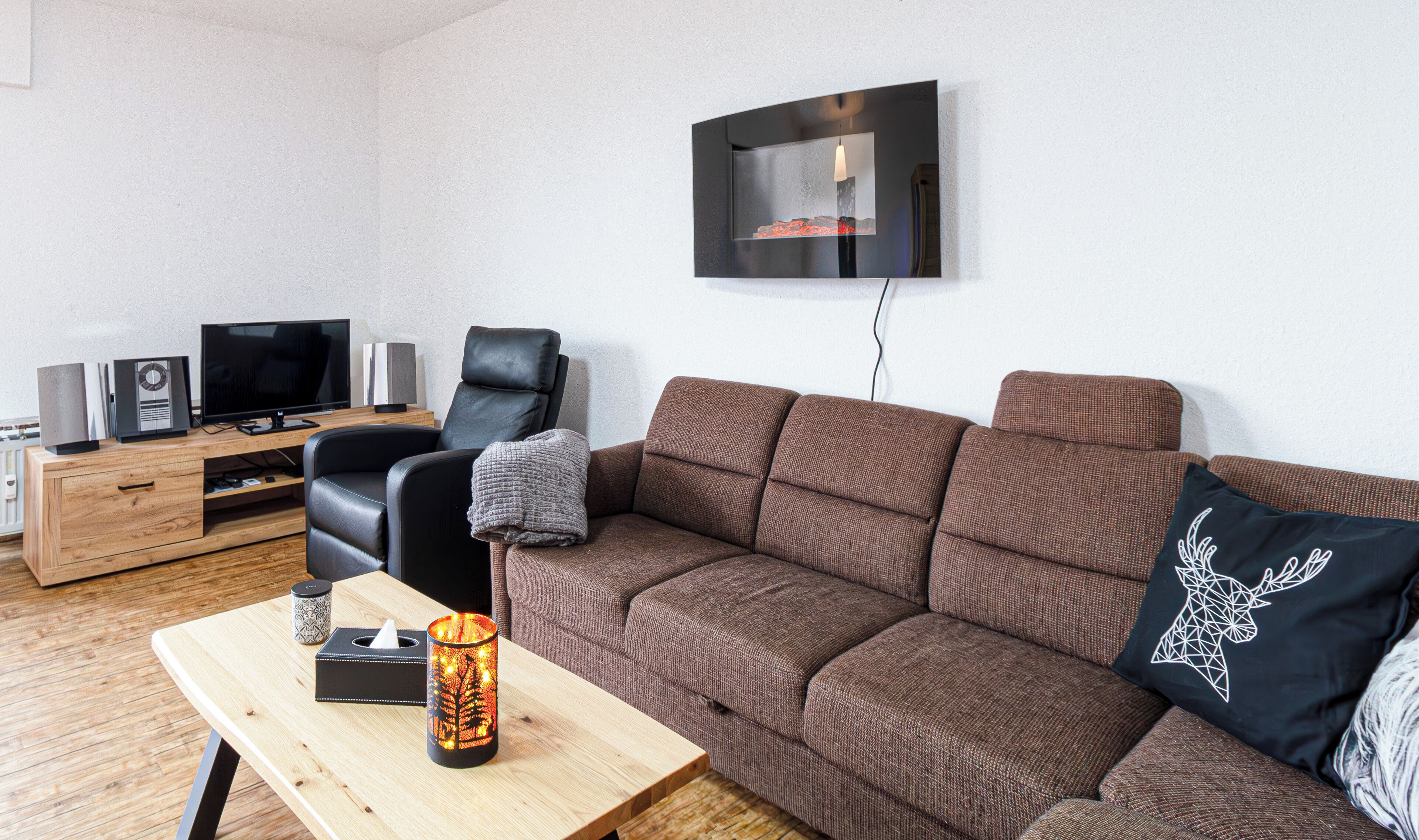 A modern living room with a brown sofa, a black leather chair, a wooden coffee table, a TV, and a mounted flat-screen TV on the wall.