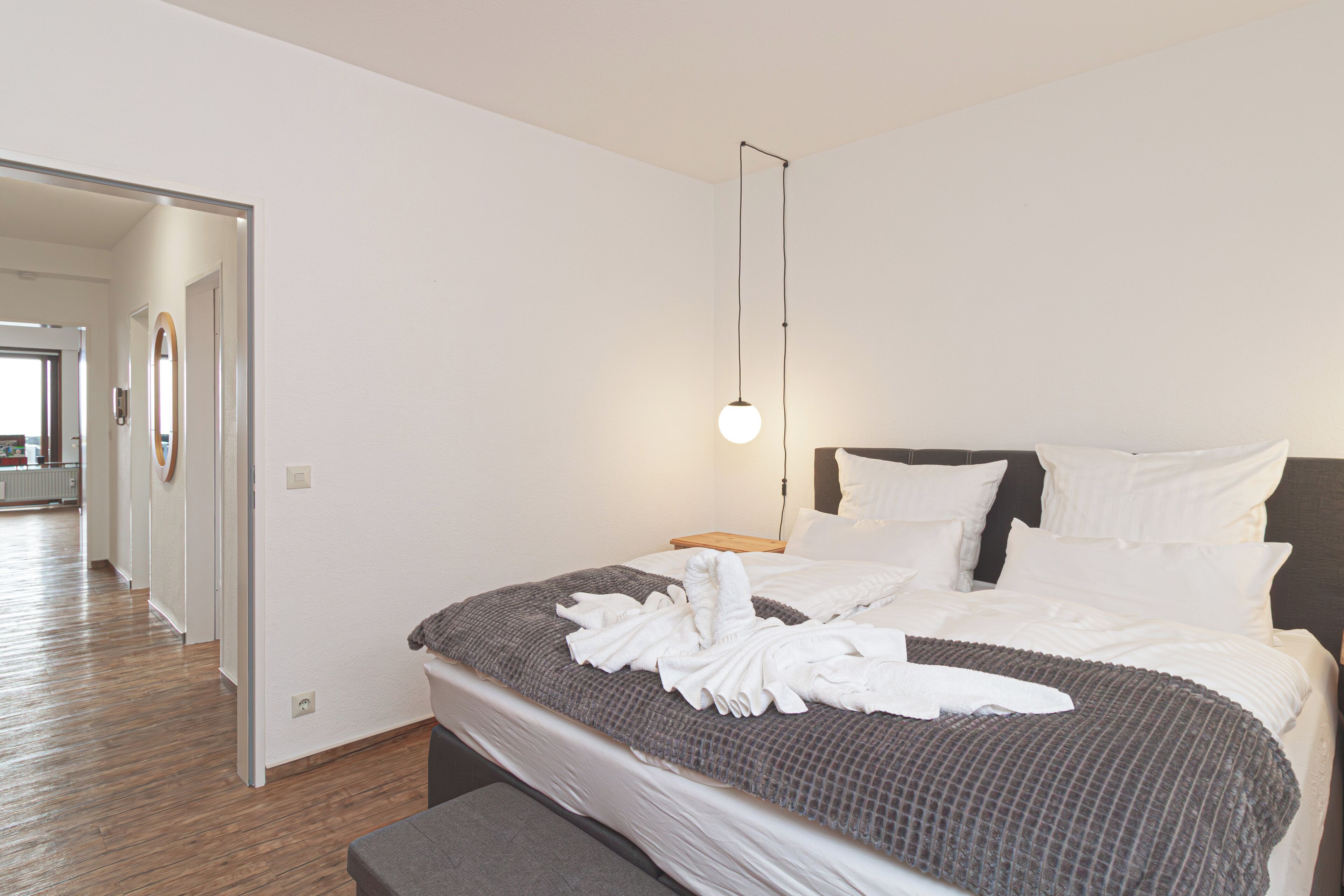 A hotel room with a neatly made bed, a bedside table, a hanging light, and a wooden floor.