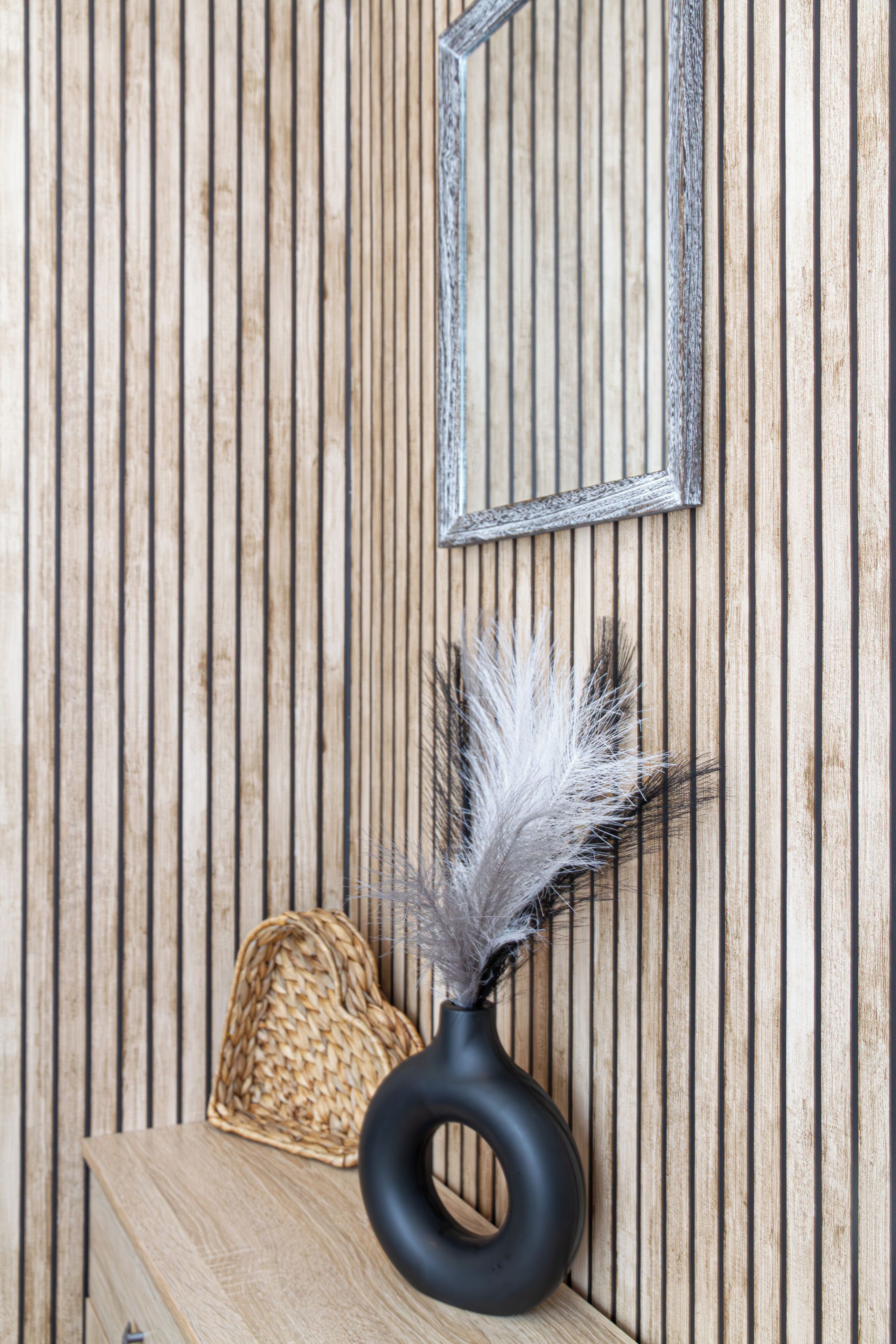 A wooden wall with vertical planks and a framed mirror. A black vase with white feathers and a woven basket are placed on a wooden surface.