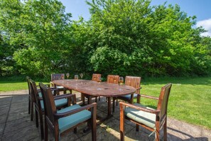 A wooden outdoor dining table with chairs, surrounded by lush greenery. - 4 Bed in Fakenham (oc-1261) (Thursford)