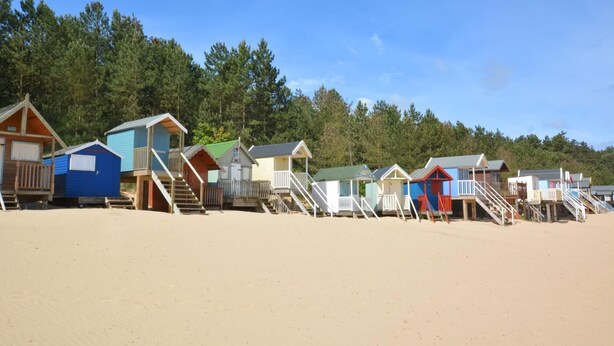 En række strandhytter med trætrapper, der fører op til dem, foran et baggrund af træer og en klar himmel.