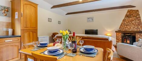 Un coin repas avec une table en bois dressée pour quatre personnes, une cheminée en briques et une cuisine dotée de meubles en bois.