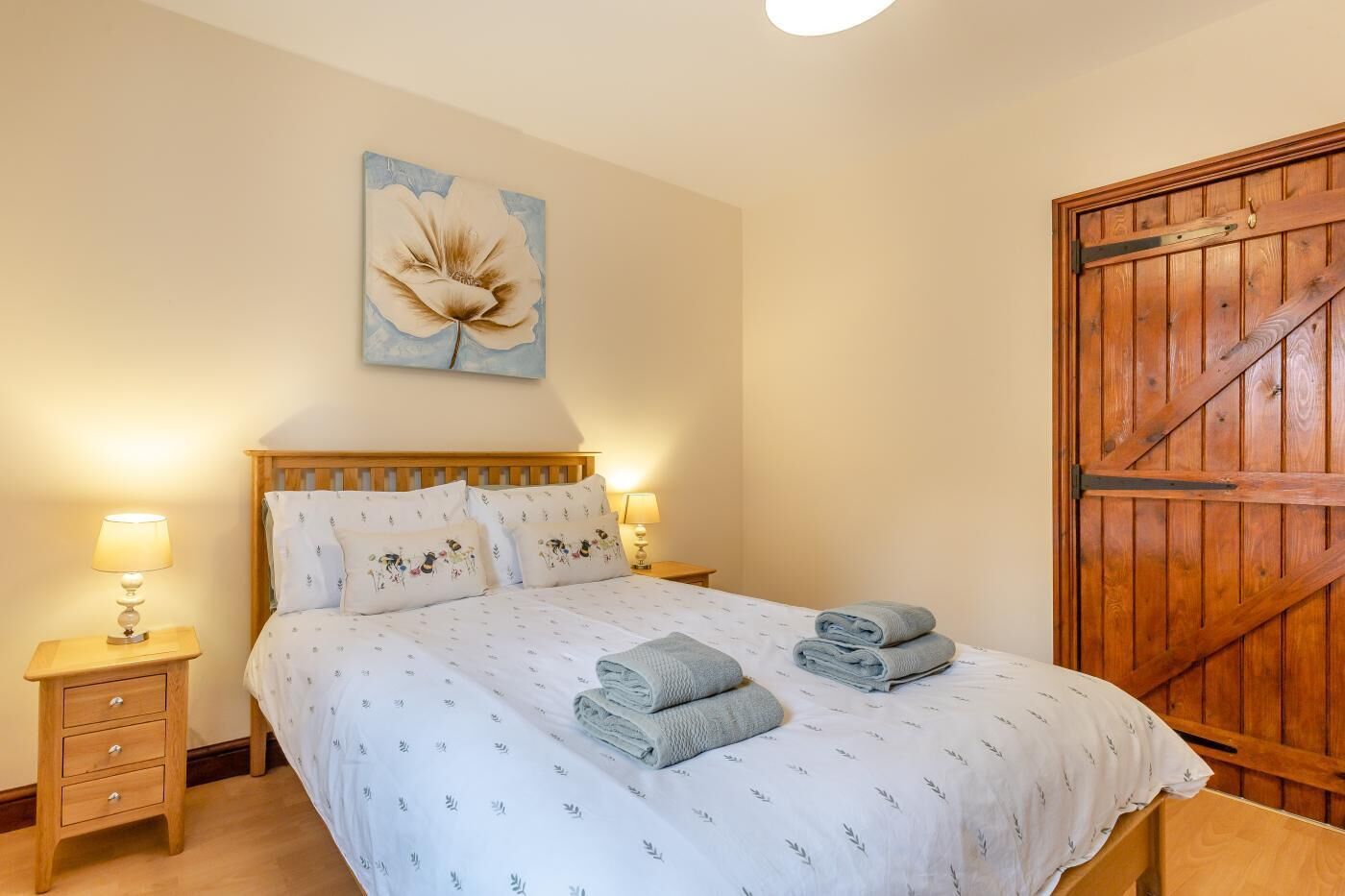 Une chambre à coucher avec un lit en bois, deux tables de chevet avec des lampes, un grand tableau floral accroché au mur et une porte en bois.