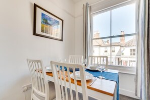 Ein Essbereich mit einem für vier Personen gedeckten Tisch, weißen Stühlen, einer blauen Tischdecke und einem gerahmten Bild an der Wand.