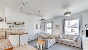 Eine moderne Küche mit weißen Schränken, einer Holzarbeitsplatte und einem eingebauten Geschirrspüler. Es gibt einen Essbereich mit weißem Tisch und Stühlen sowie ein Wohnzimmer mit einem beigefarbenen Sofa und einem kleinen Holztisch. Große Fenster sorgen für natürliches Licht und Ausblicke nach draußen.