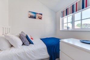 Une chambre à coucher avec un lit, des oreillers, une couverture bleue, une fenêtre avec des rideaux à rayures et deux tableaux accrochés au mur.