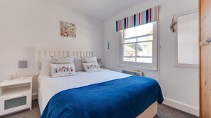 Ein Schlafzimmer mit einem Bett, weißen Nachttischen, einem mit Vorhängen drapierten Fenster und einem wandmontierten Spiegel.