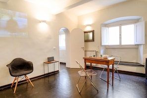 Ein Zimmer mit dunklem Holzboden, einem schwarzen Stuhl, einem kleinen Tisch mit einem Laptop, einem transparenten Stuhl und einem Fenster mit weißen Läden.