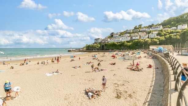 Ein Strand mit badenden und sonnenden Personen, eine Promenade mit einer Brüstung und Häuser, die sich einen Hügel hinaufziehen.