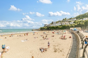 Ein Strand mit badenden und sonnenden Personen, eine Promenade mit einer Brüstung und Häuser, die sich einen Hügel hinaufziehen.
