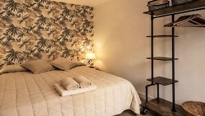 Ein Bett mit einer gesteppten Decke, eine Wand mit tropischem Blattmuster, ein Nachttisch mit Lampe und ein Metallregal mit Körben.
