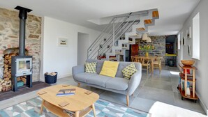 A modern living room with a stone fireplace, a grey sofa with yellow cushions, a wooden coffee table, and a staircase leading to an upper level. - 3 Bed in Botwnnog (oc-wag571) (Botwnnog)
