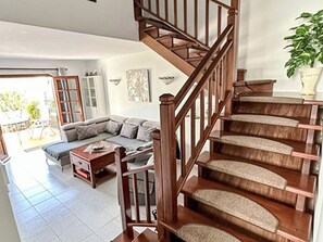 Eine Treppe mit Holzwerteln führt zu einer oberen Etage, dort befindet sich ein graues Ecksofa, ein Holztisch und ein Blumentopf.