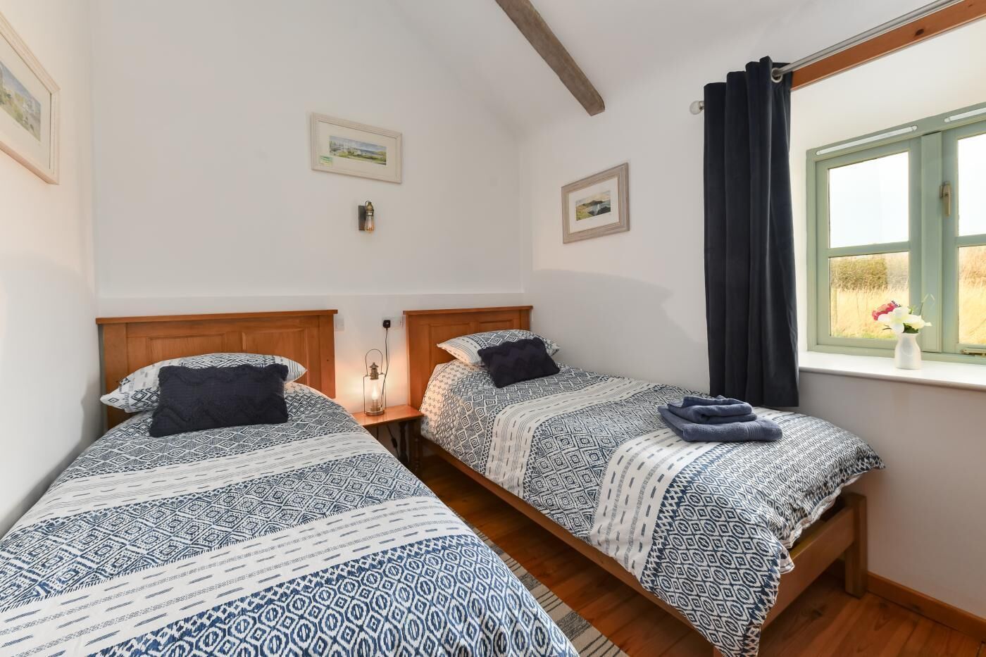 A room with two beds, wooden headboards, a window with curtains, and a small table with a lamp.