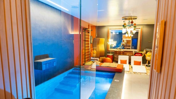 A modern hotel room with a glass-enclosed pool, a seating area with orange cushions, and a large mirror reflecting the room.