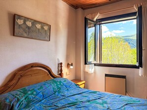 Ein Schlafzimmer mit einem Bett, einem Nachttisch, einer Lampe, einem Fenster mit Blick auf Bäume und einem Bild an der Wand.