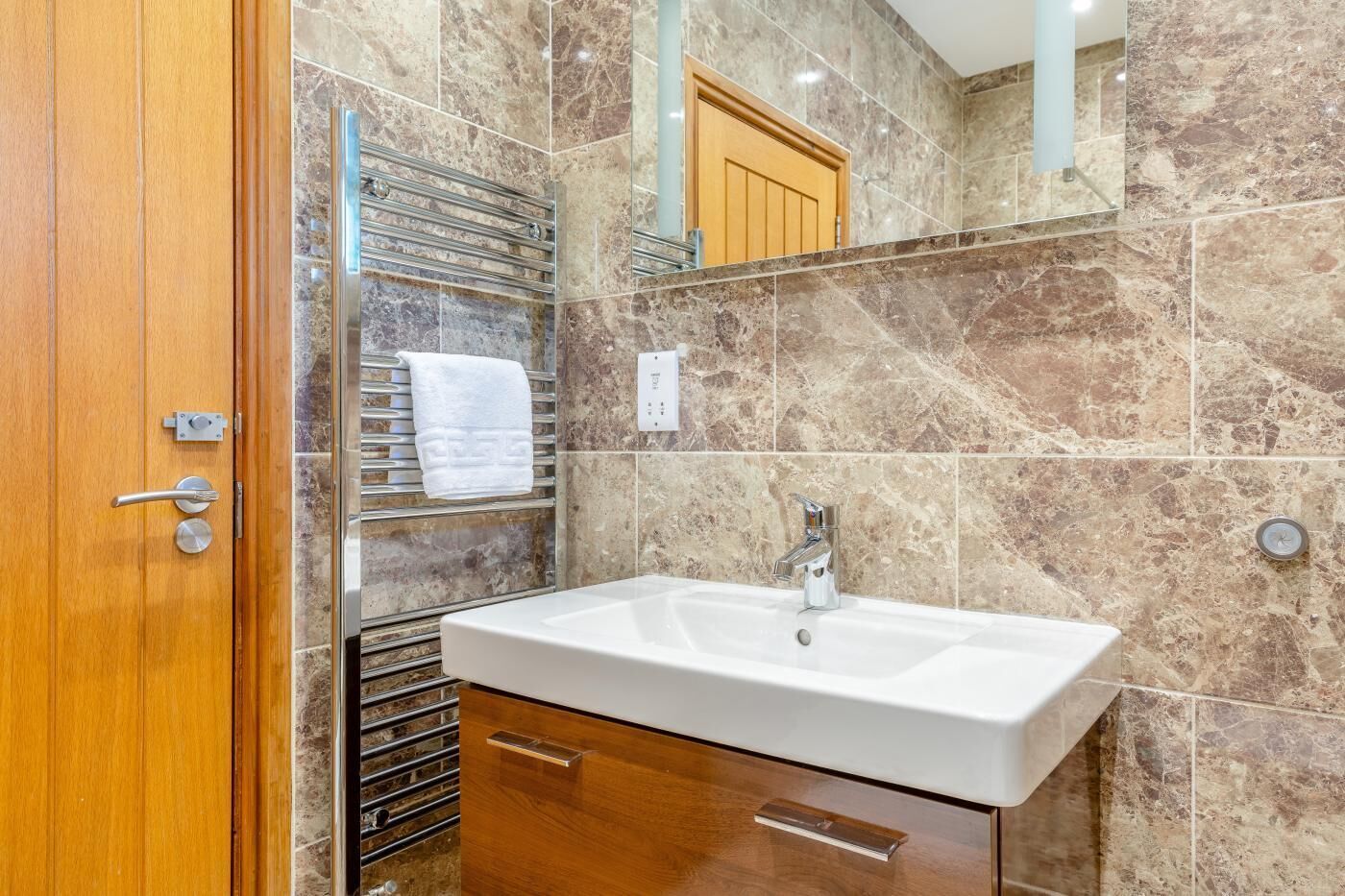 Une salle de bain avec une porte en bois, un lavabo blanc avec un robinet chromé et un porte-serviettes sur lequel est posée une serviette blanche.