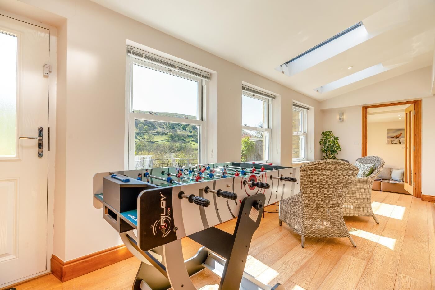 A room with a foosball table, large windows, a wicker chair, and a view of greenery outside.
