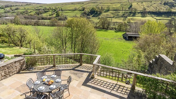 Un patio con una mesa lista para comer, con vista a un paisaje verde con colinas y un edificio pequeño.